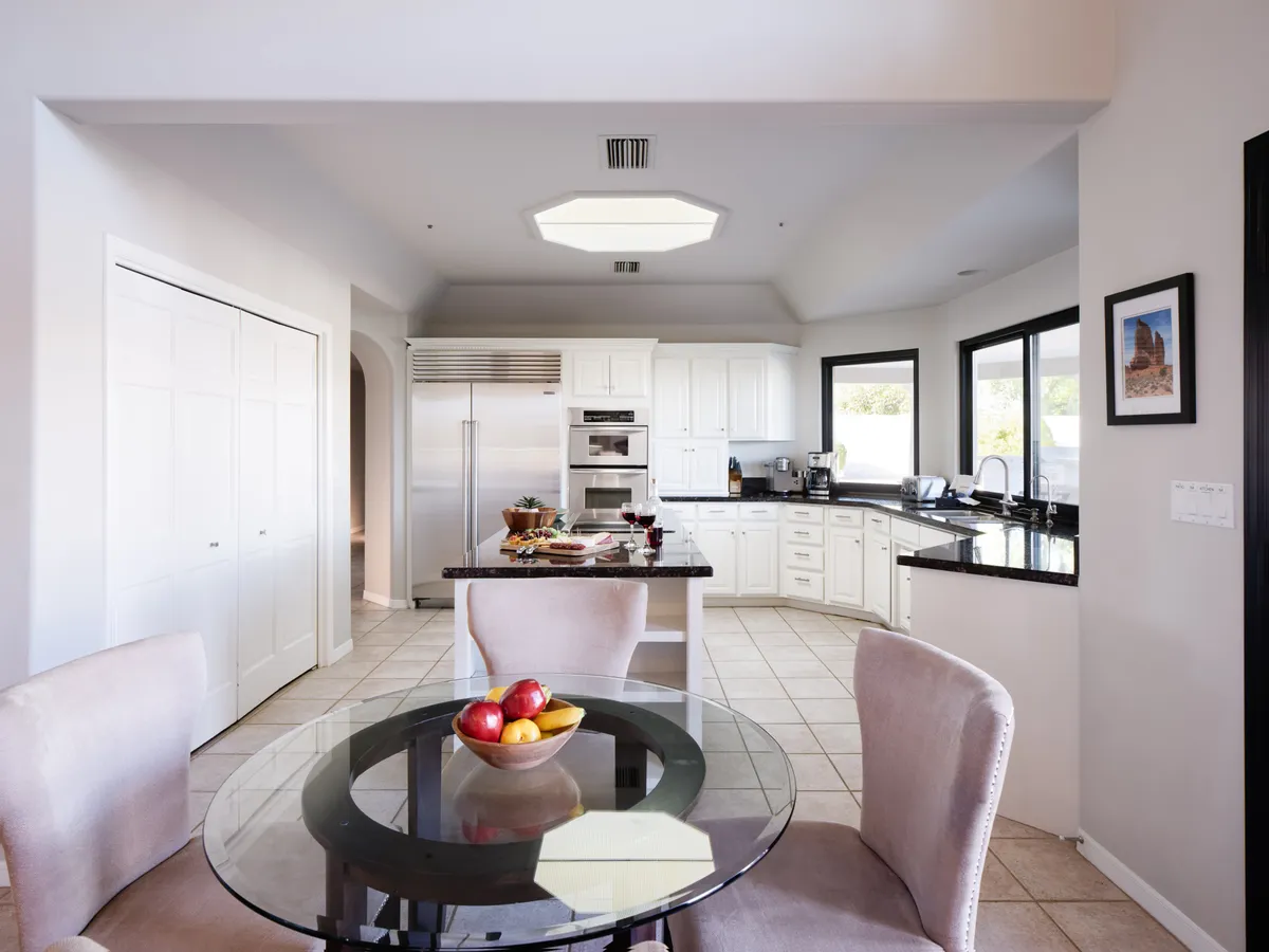 A modern kitchen with sleek white cabinetry, stainless steel appliances, and a glass table set with a fruit bowl near large windows. The space is bright and welcoming, ideal for hosting, morning rituals, or casual get-togethers.