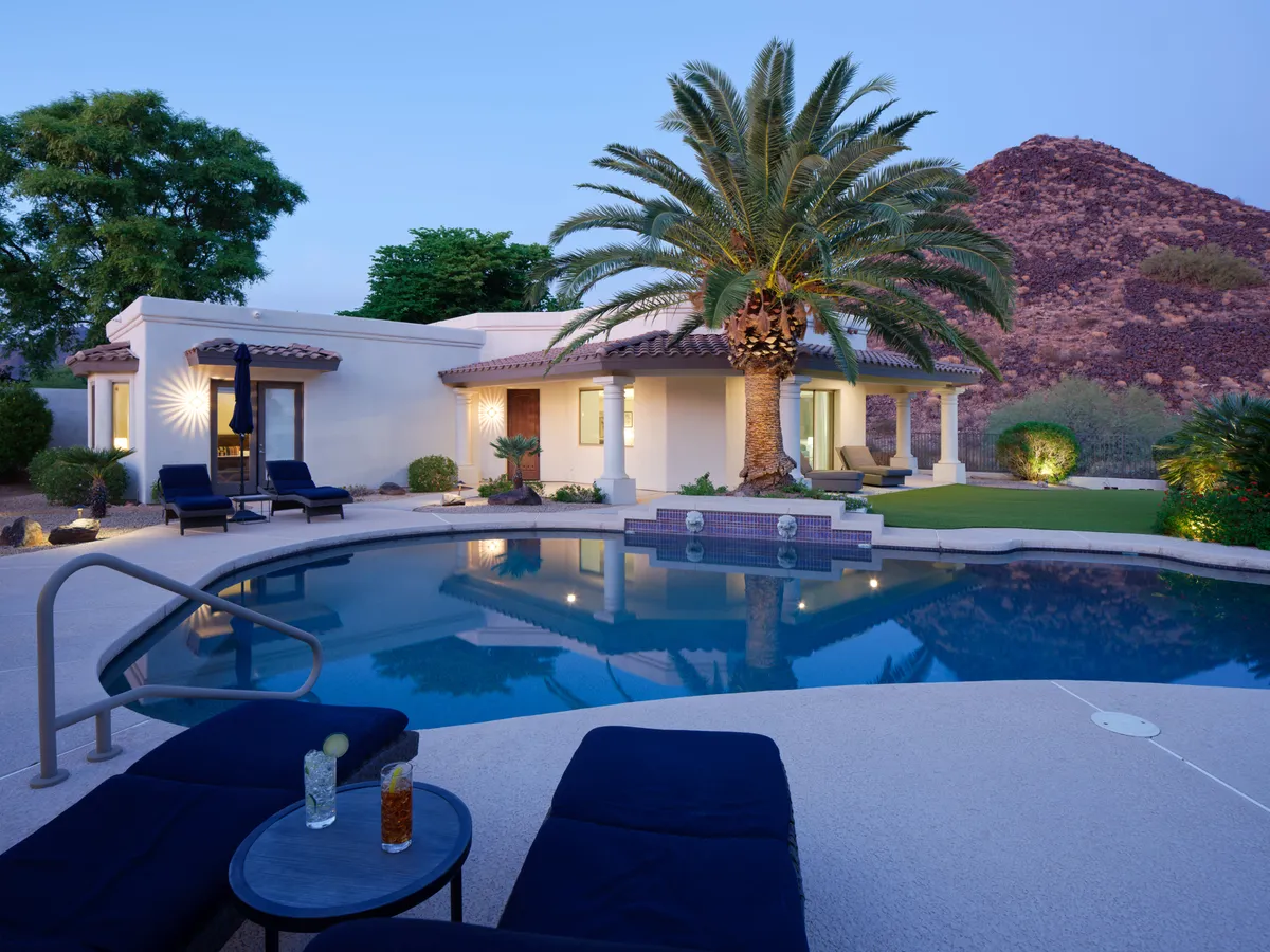 The hero image showcases the estate's sparkling pool surrounded by elegant loungers and swaying palm trees, set against a dramatic mountain backdrop. The scene glows with warm sunset light, hinting at relaxing swims, poolside cocktails, and the natural beauty of the Arizona desert.