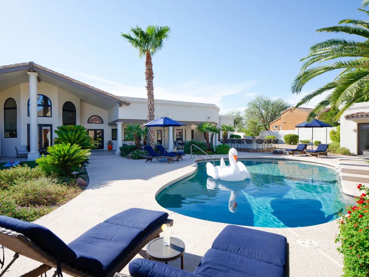 A luxurious backyard scene with a swimming pool, a whimsical swan float, sun loungers shaded by umbrellas, and tall palm trees. The overall ambiance is one of relaxed luxury, inviting guests to swim, sunbathe, or simply unwind in a beautiful setting.