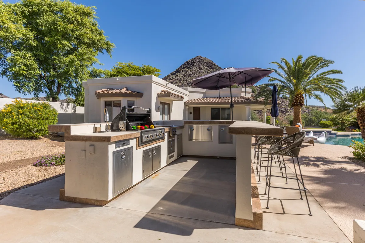 A stylish outdoor kitchen and grill area with bar stools, shaded by palm trees and set against a mountain backdrop. The kitchen overlooks the pool and provides plenty of space for cooking, dining, and gathering with friends or family.