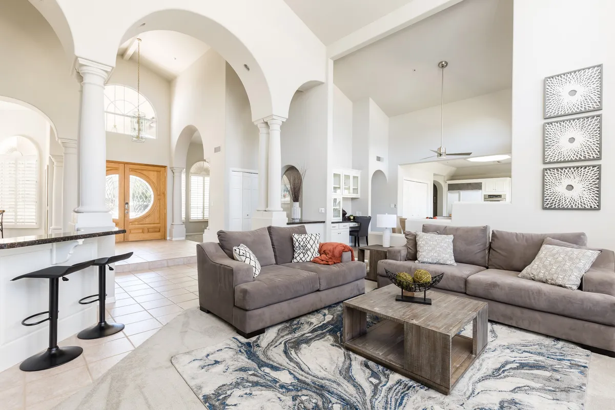 The image showcases a spacious living room bathed in natural light, featuring comfortable sofas with decorative pillows, a coffee table with a stylish centerpiece, and a cozy area rug. Large windows create an airy, welcoming atmosphere ideal for socializing or relaxing, perfectly reflecting the estate's main living area.