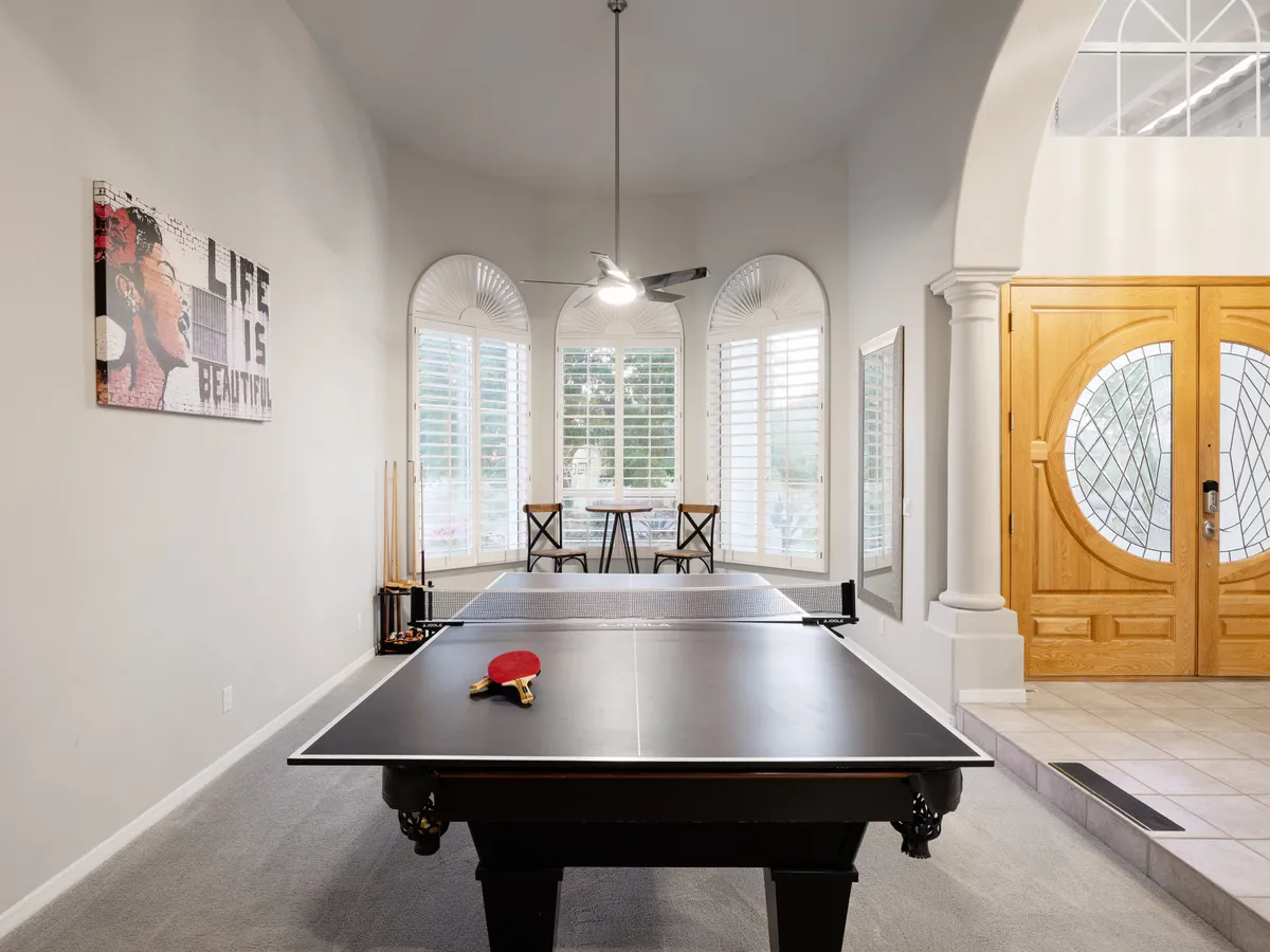 A welcoming interior scene with a modern ping pong table set up in the foreground, decorative artwork on the walls, and a visible dining area with chairs. Ornate entrance doors add flair to the space, which feels open, engaging, and ideal for group activities.