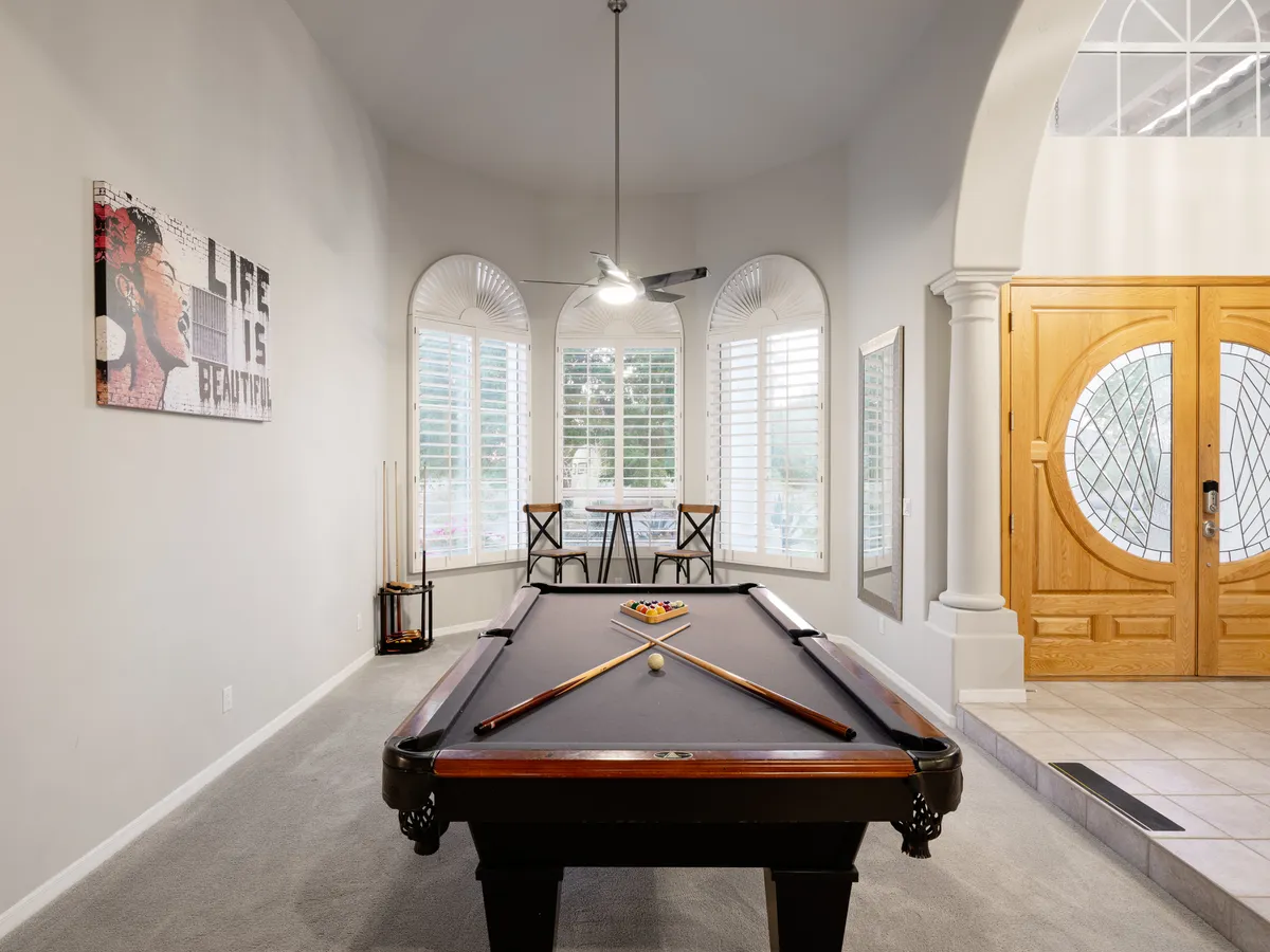 This image presents a vibrant game room featuring a pool table as the focal point, complemented by large wooden doors, a dining table with chairs, and stylish wall art. The space is well-lit and arranged for both competitive play and relaxed social gatherings.