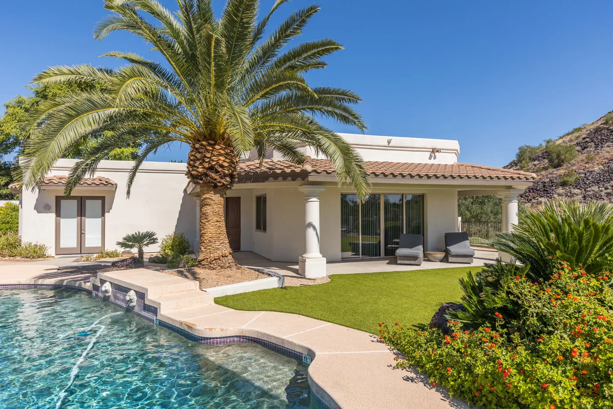 A tranquil backyard scene featuring a sparkling swimming pool surrounded by lush palm trees and a manicured lawn. The house's modern facade frames an inviting patio area ideal for dining or relaxation, while the setting sun casts a warm glow over the space.