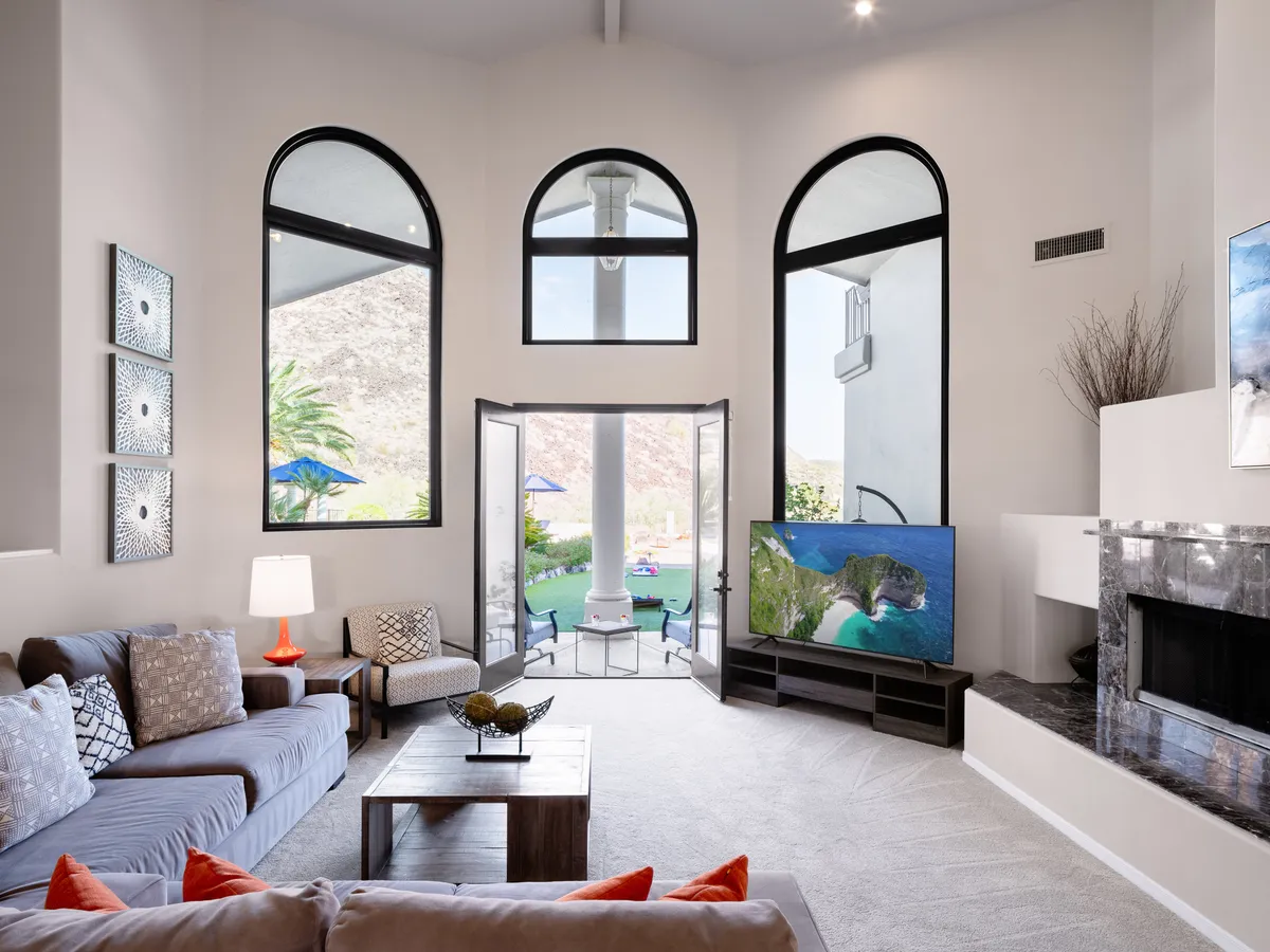 A comfortable seating area with large windows showcasing the outdoors, decorative pillows, double doors leading outside, and a modern television. The arrangement encourages group relaxation, conversation, or movie nights, exemplifying the welcoming spirit of the estate's gathering spaces.