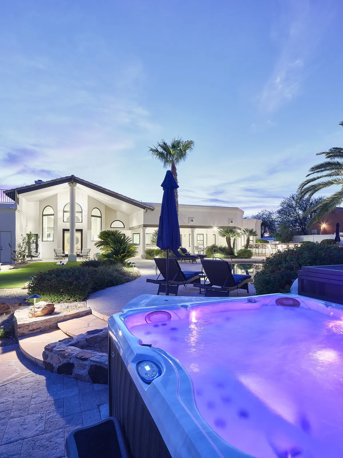 A luxurious outdoor area at dusk, highlighting a hot tub with vibrant purple lighting. Comfortable seating and palm trees surround the space, and the modern architecture of the home is visible in the background, creating an inviting spot for evening relaxation under the stars.