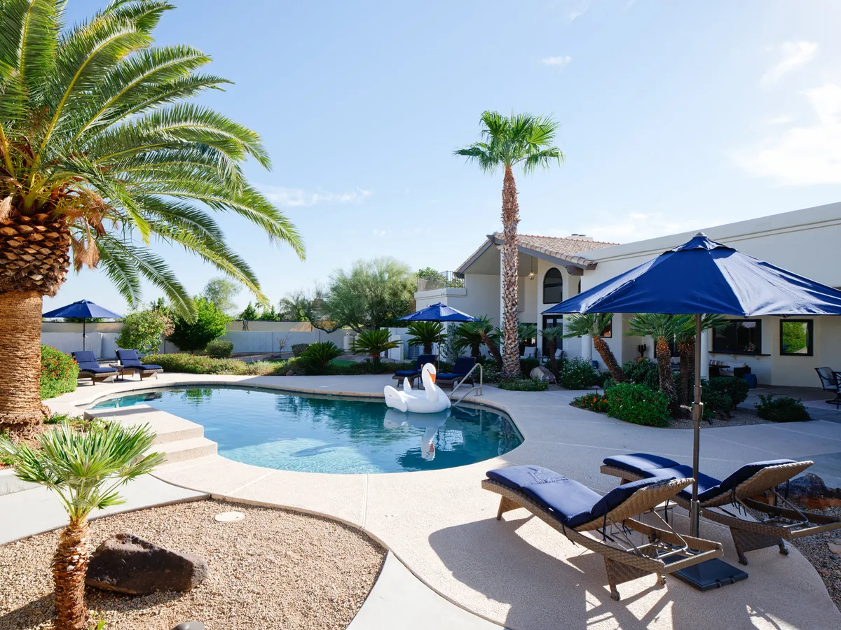 A cheerful backyard pool scene with a swan float, blue umbrellas, palm trees, and plenty of lounge chairs. Children are visible playing, capturing the fun, family-oriented atmosphere of poolside afternoons.