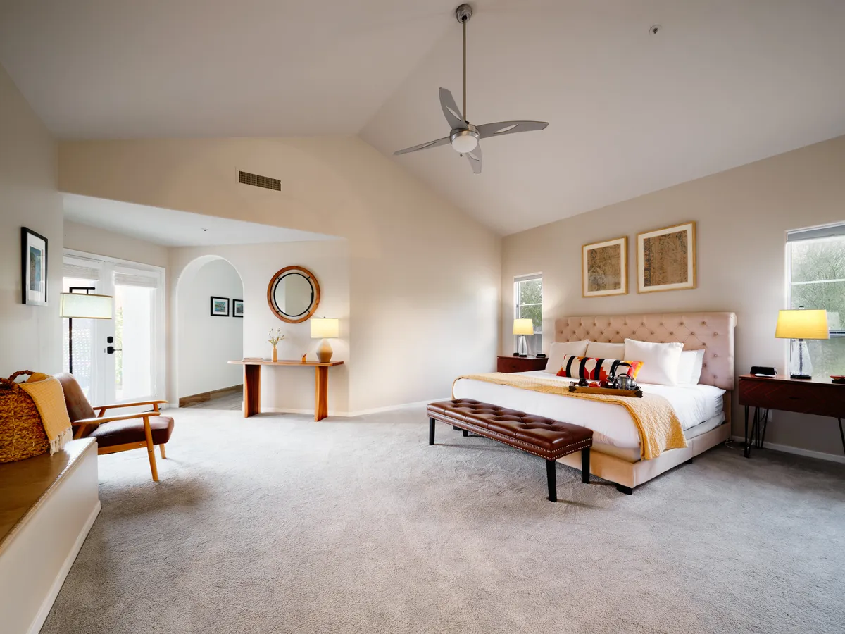 This image shows a serene bedroom with a king-sized bed, a bench at the foot, an armchair, a work desk with a lamp, and tasteful decor. Natural light flows in, highlighting the calm, cozy atmosphere.