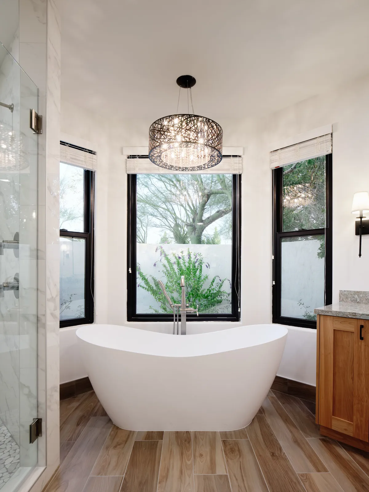 A serene interior bathroom featuring a freestanding bathtub beneath a sparkling chandelier, with large windows offering a view of greenery and elegant wooden cabinetry. The space exudes tranquility and luxury, ideal for relaxation and self-care routines.